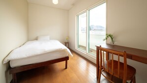 A bedroom with a wooden bed, a small bedside table with a vase, a wooden desk with a chair, and a large window with a view of buildings and hills. - Cafe&Guest House Kaziya (Ichinoseki)
