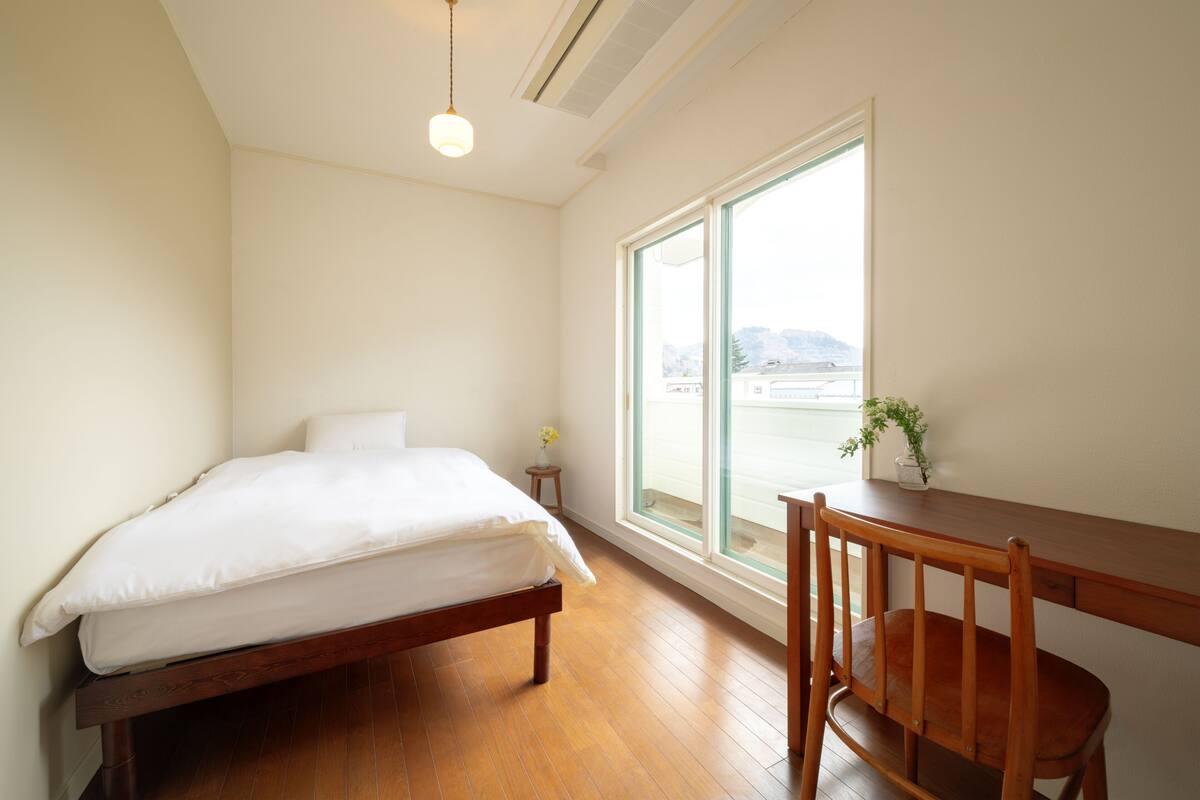 A bedroom with a wooden bed, a small bedside table with a vase, a wooden desk with a chair, and a large window with a view of buildings and hills.