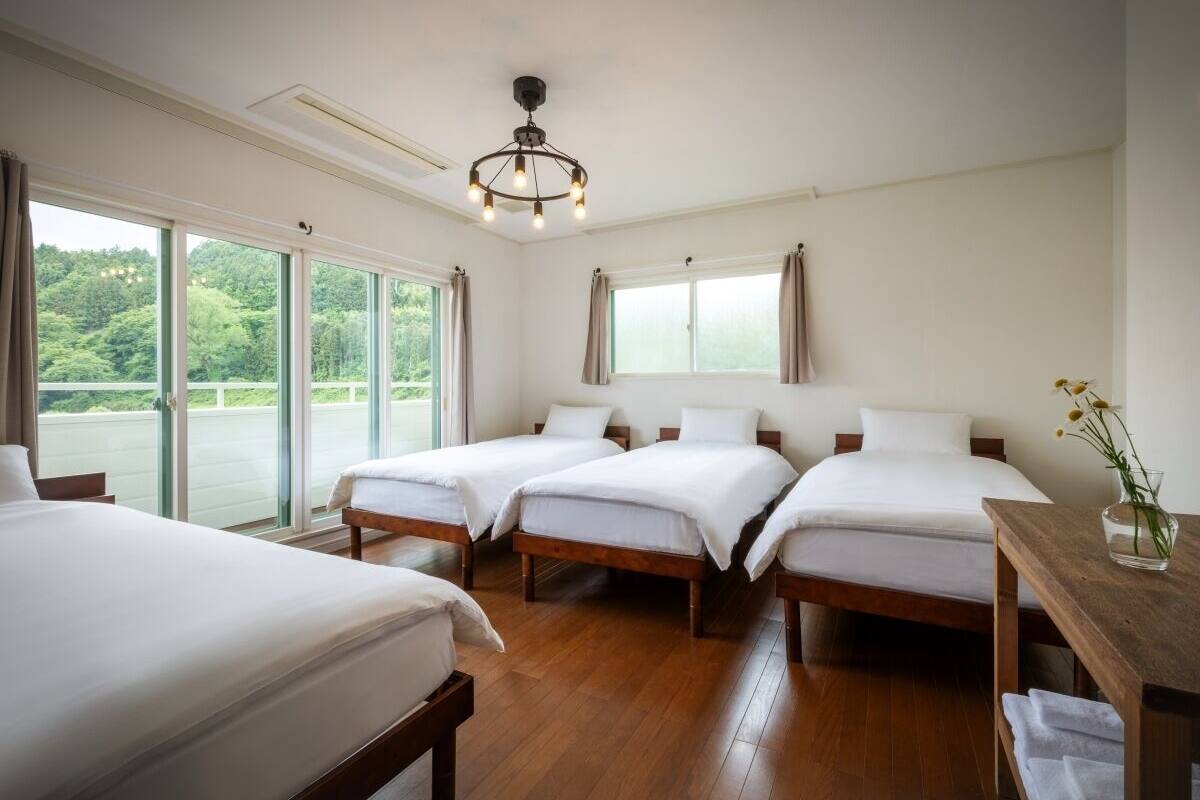 A hotel room with four beds, a wooden floor, a chandelier, and large windows with curtains.