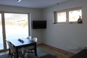 A room with a dining table, chairs, a flat-screen TV, a window with a view of mountains, and a small shelf with decorative items.