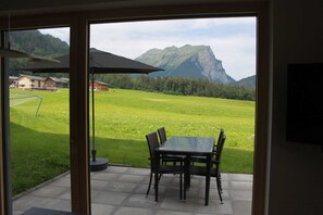 A patio with a table and chairs, a large umbrella, and a view of a mountain and buildings. - Kanistraum apartment (Schoppernau)