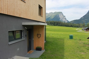 A modern building with a wooden facade, a small entrance with a heart-shaped decoration, and a view of a green field and mountains in the background. - Kanistraum apartment (Schoppernau)