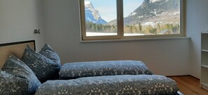 A bedroom with a bed, patterned pillows, a window with a mountain view, and a wooden floor. - Kanistraum apartment (Schoppernau)
