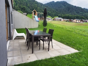 Un patio con una mesa de vidrio y sillas de mimbre, un parque infantil y vistas a una ladera verde.