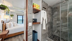 Une chambre d’hôtel comprenant un lit, une table de chevet, une commode, une salle de bain avec douche, un porte-serviettes et une plante.