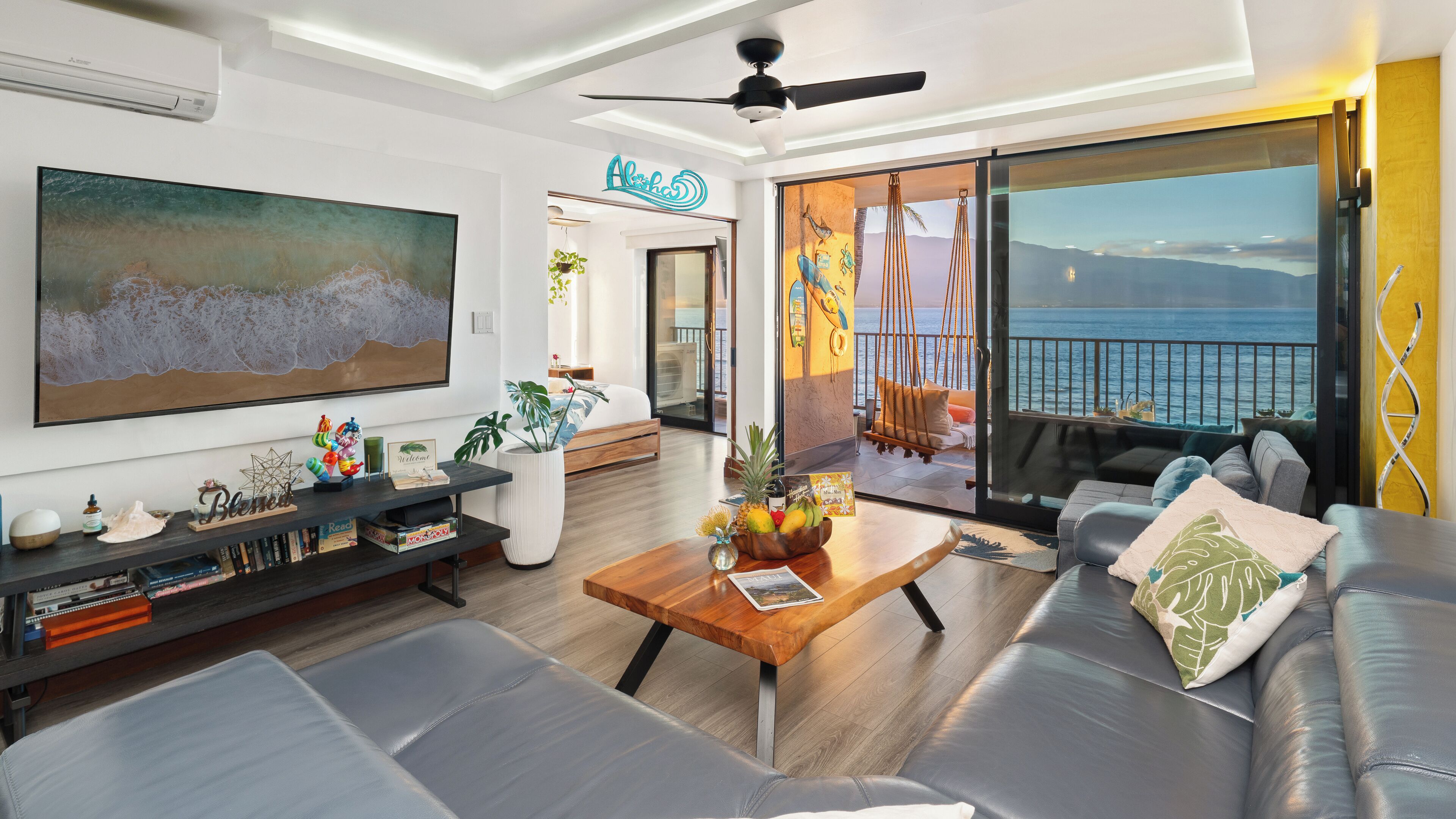 A modern living room with a large painting, a sofa, a coffee table with fruit, and a view of the ocean.