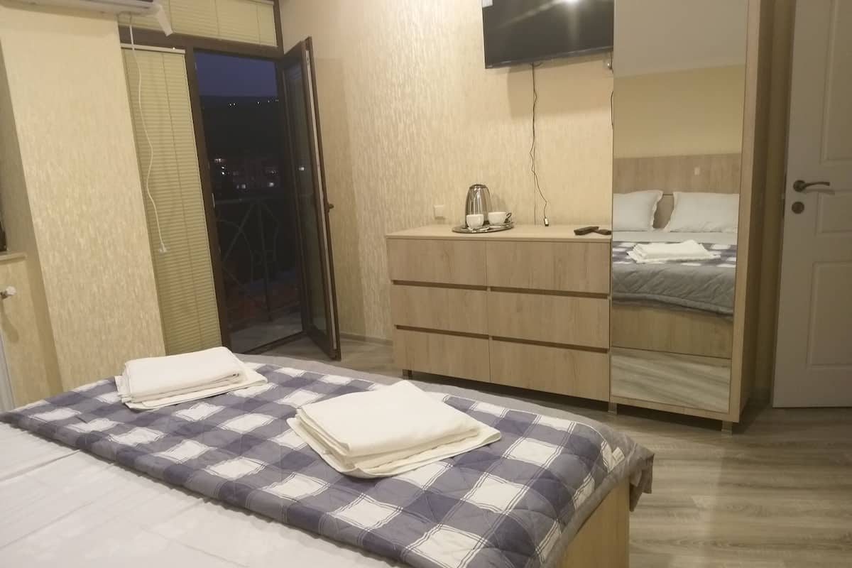 A bedroom with a bed, a wooden dresser, a TV mounted on the wall, and a sliding door leading to a balcony.