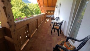 Un balcón con barandilla de madera, una mesa con sillas y vistas a zonas verdes y montañas.
