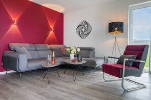 Una sala de estar moderna con un sofá gris, un sillón rojo y una mesa de centro. Hay un adorno en la pared y una lámpara de pie.