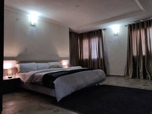 Una habitación de hotel con una cama grande, dos lámparas de noche, un tapete oscuro y cortinas en las ventanas.