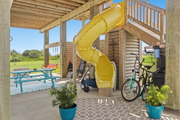 A wooden play structure with a yellow slide, picnic tables, and potted plants.