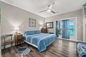 A bedroom with a bed, a chair, a nightstand, a lamp, a ceiling fan, and a view of the outside.