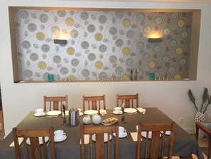 Ein Essbereich mit einem Tisch, der für sechs Personen gedeckt ist, umgeben von Holzstühlen. An den Wänden sind Leuchten angebracht und eine Wand ist mit einem floralen Muster verziert.