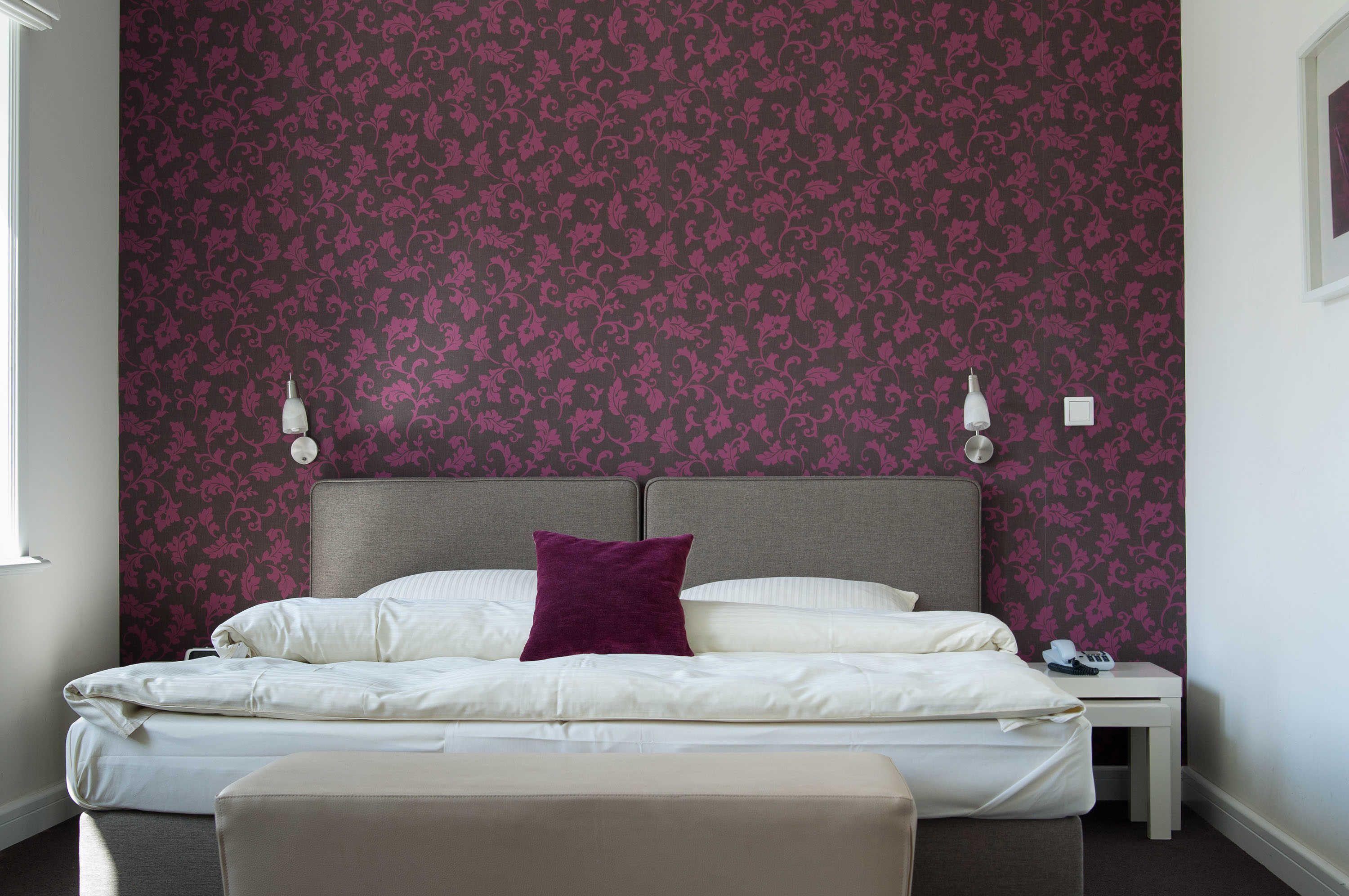 A neatly made bed with a headboard, a bedside table with a lamp, and a wall with a floral pattern.