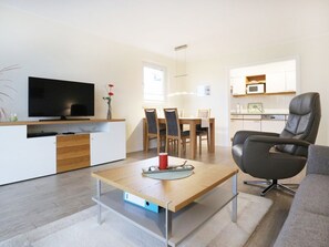 Una sala moderna con televisor de pantalla plana, comedor con sillas, cocina con microondas y un sillón de cuero.
