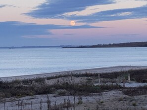 Una playa con un mar tranquilo, una luna visible y una costa distante.
