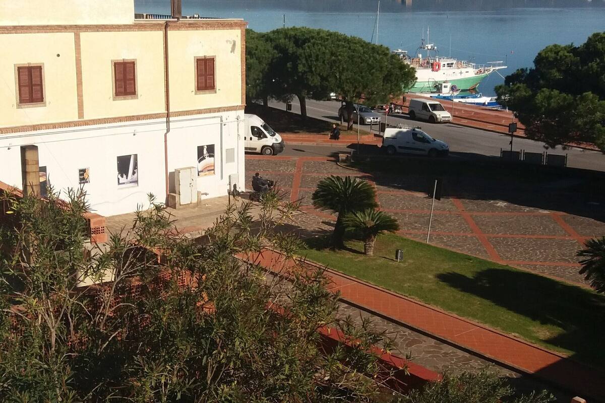 Vista costera con un edificio, coches aparcados y un muelle para barcos.