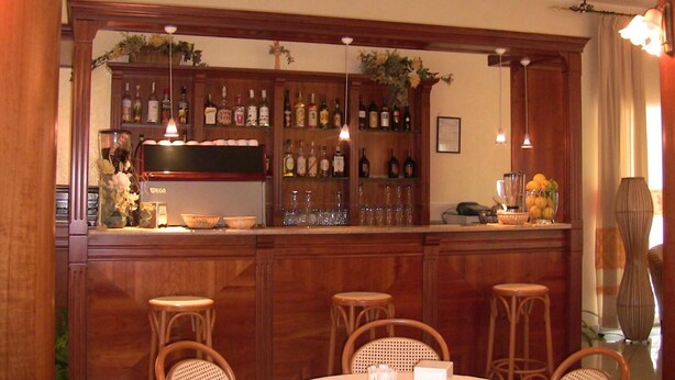 Zona de bar con armarios de madera, botellas, vasos y sillas de mimbre.