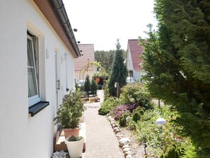 A narrow pathway flanked by white buildings with red-tiled roofs, leading to a green area with potted plants and a small garden. - Bungalow Vincent, with terrace, barbecue & WLAN - Bungalow with garden, terrace and (Zempin)