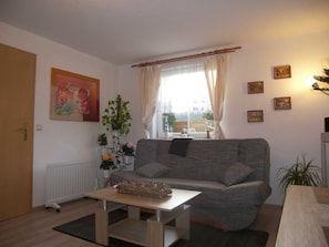 Un salon avec un canapé gris, une table basse, un radiateur, une fenêtre avec des rideaux et plusieurs tableaux encadrés accrochés au mur.