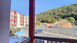 Vista desde un balcón sobre un estacionamiento con varios coches, un edificio de fachadas rojas y beige, y una ladera boscosa al fondo.