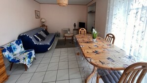 Ett vardagsrum med en soffa, ett matbord och stolar. Det står en lampa på ett sidobord, en element och en tv på ett TV-bord.