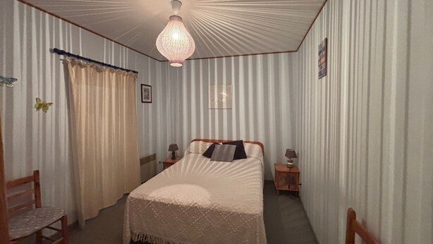 Ett sovrum med en säng, två sängbord, en trästol, en hängande lampa och ett fönster med gardiner.