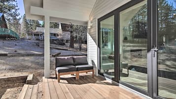 Terraza de madera con un sofá gris, un ventanal de vidrio que da a un patio, y vistas a un jardín trasero con otra terraza de madera y una casa al fondo.