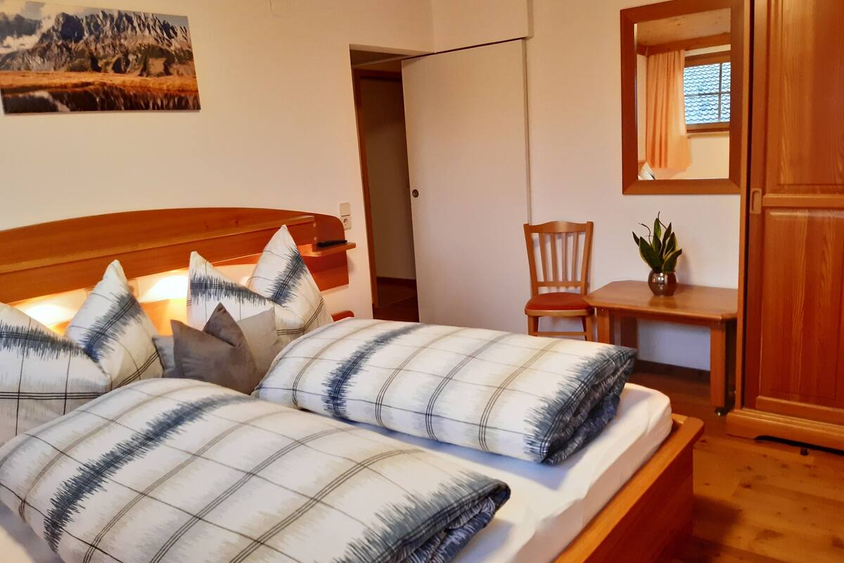 A bedroom with a bed, a chair, a small table, and a potted plant.