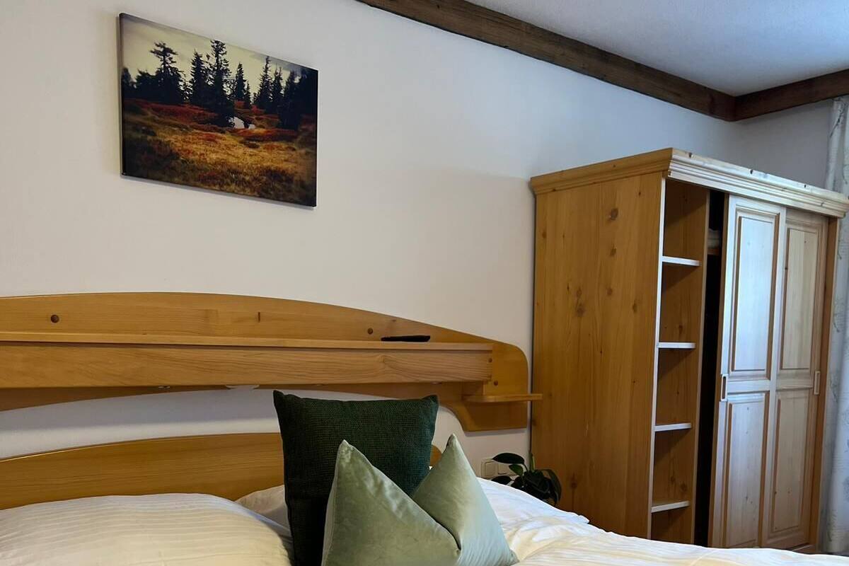 A double bed with a wooden headboard, two pillows, and a painting of a forest on the wall.