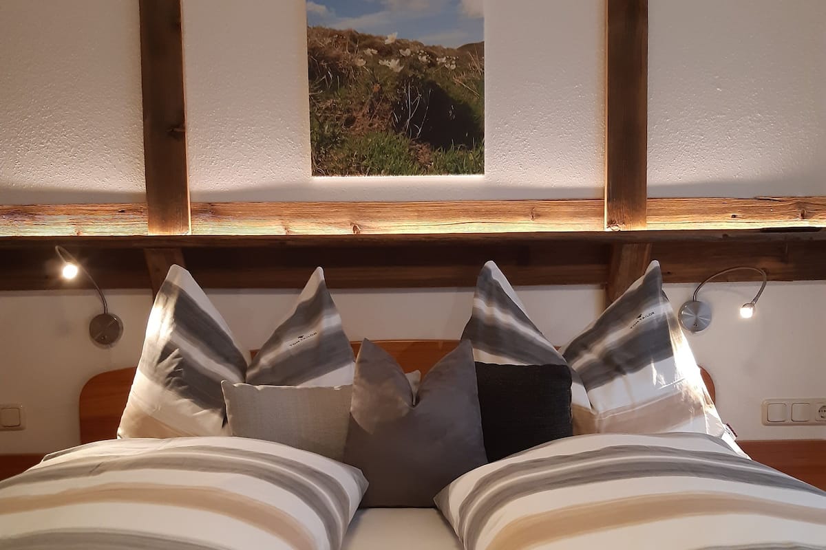 A bed with striped bedding and multiple pillows, a wooden headboard, a small window with a view of a landscape, and wall-mounted lights.