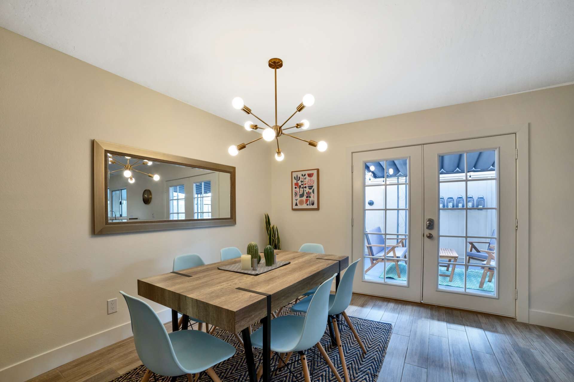 A dining area with a wooden table, blue chairs, a chandelier, a large mirror, and a door leading to a balcony with chairs.