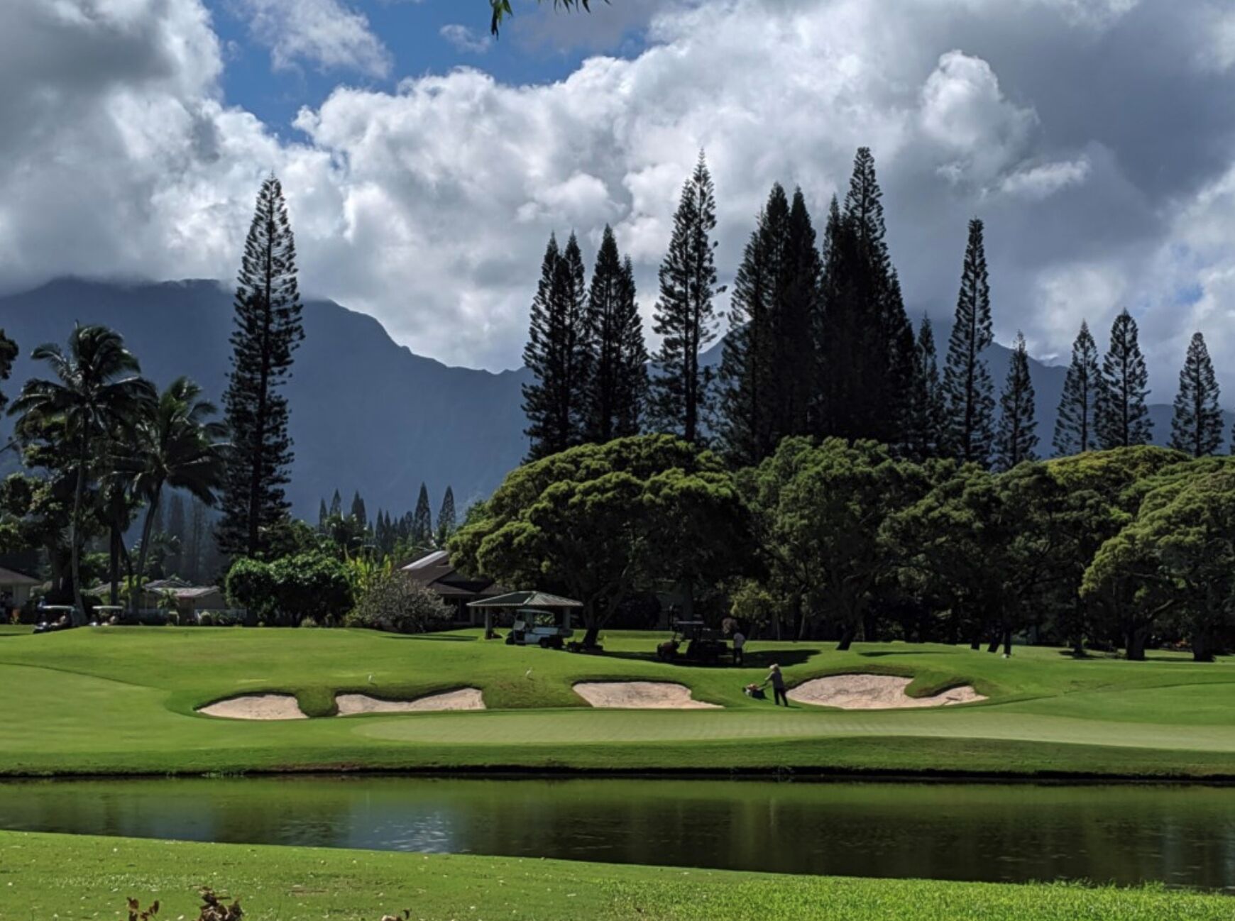 Ein Golfplatz mit Sandbunkern, einem Teich und einer Kulisse aus hohen Bäumen und Bergen.