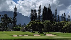 A golf course with sand bunkers, a pond, and a backdrop of tall trees and mountains. - North Shore Kauai- AC, pool, lanai; near beaches, golf, walking path to shops (Princeville)