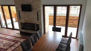 A dining area with a long wooden table, black cushioned chairs, a patterned rug, and a stone fireplace. - Meshtekna (Brezovicë)