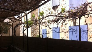 A covered outdoor area with a stone wall, a pergola, and a view of a building with blue shutters. - Character house at the foot of the castle. Shaded terrace (Boulbon)