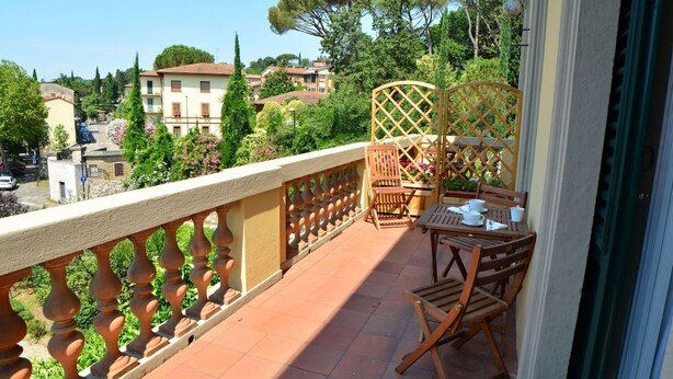 Un balcon avec une table et des chaises en bois, un fauteuil en osier et une vue sur un quartier résidentiel, avec des arbres et des immeubles.