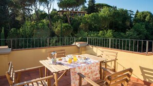 A terrace with a table set for breakfast, surrounded by wooden chairs, overlooking a lush garden and a building in the background. - Apartment with terrace ovelooking Florence (Firenze)