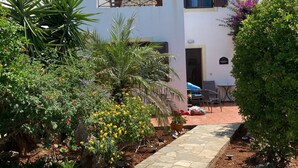 Ein Weg führt zu einem Gebäude mit einer Pergola, umgeben von Pflanzen und einer Terrasse mit Gartenmöbeln.