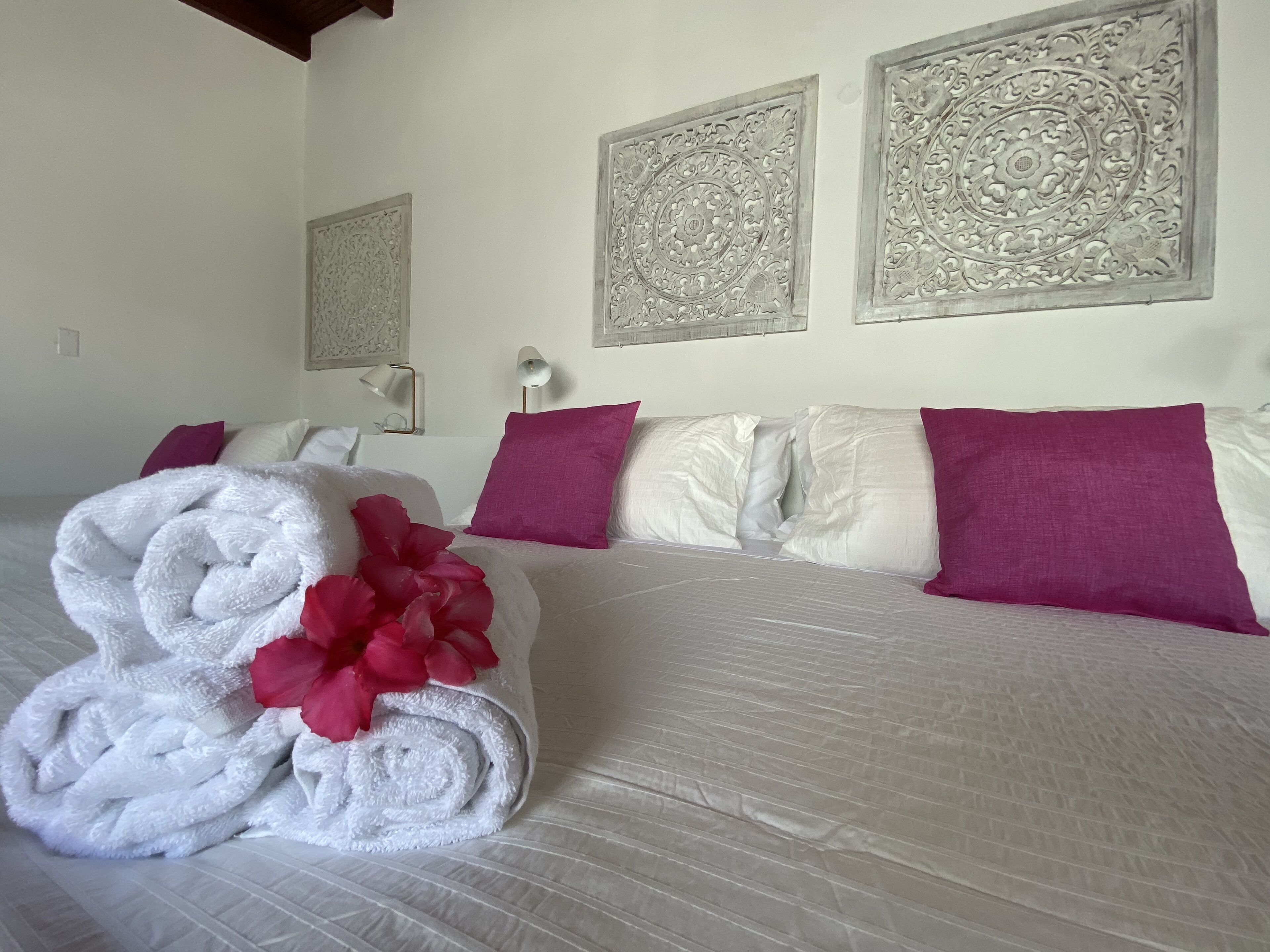 A neatly made bed with white linens and a pink flower, two purple pillows, and decorative wall panels.