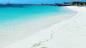 Una playa con agua turquesa cristalina, arena blanca y un velero a lo lejos.