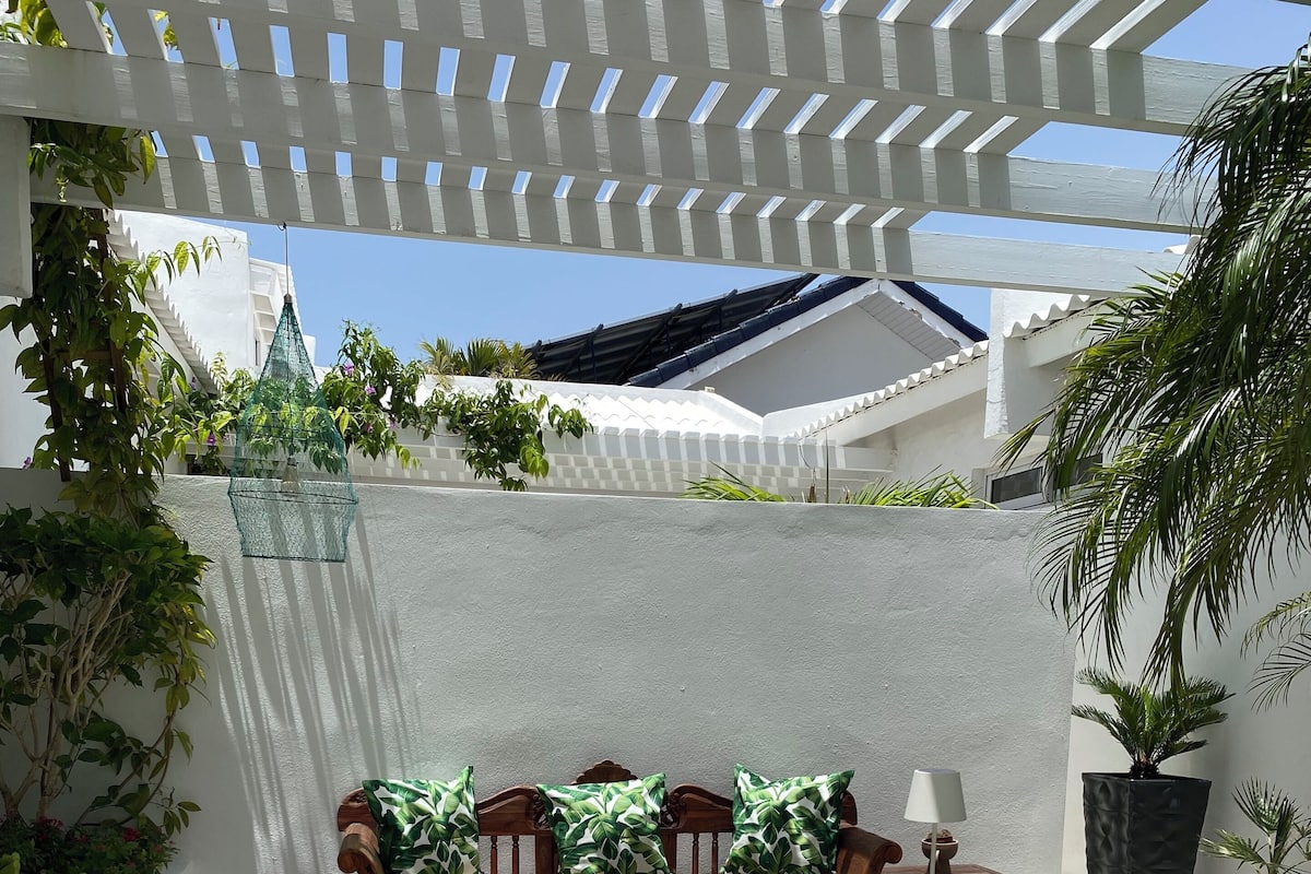 A patio with a white pergola, a wooden sofa with green cushions, a small table, and potted plants.