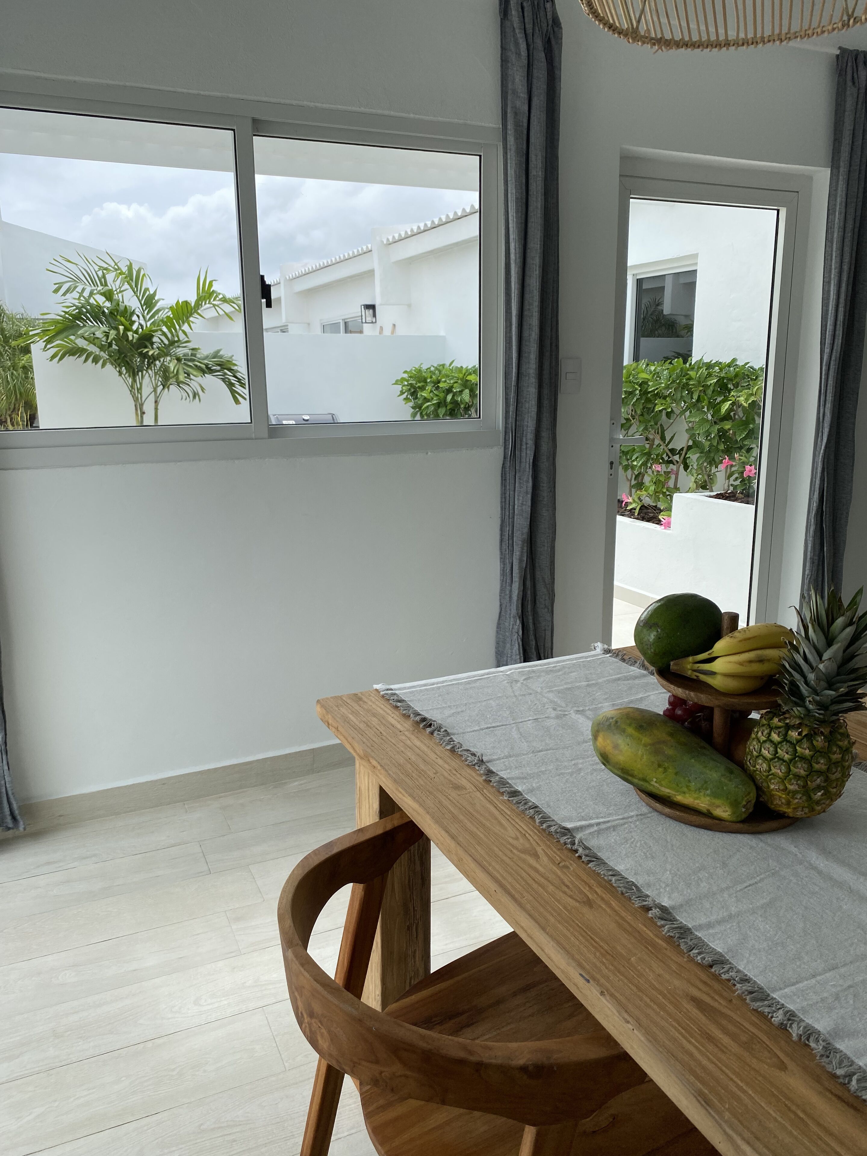 A wooden dining table with a grey tablecloth, a fruit bowl with pineapple, banana, and mango, and a view of a white building and plants outside.