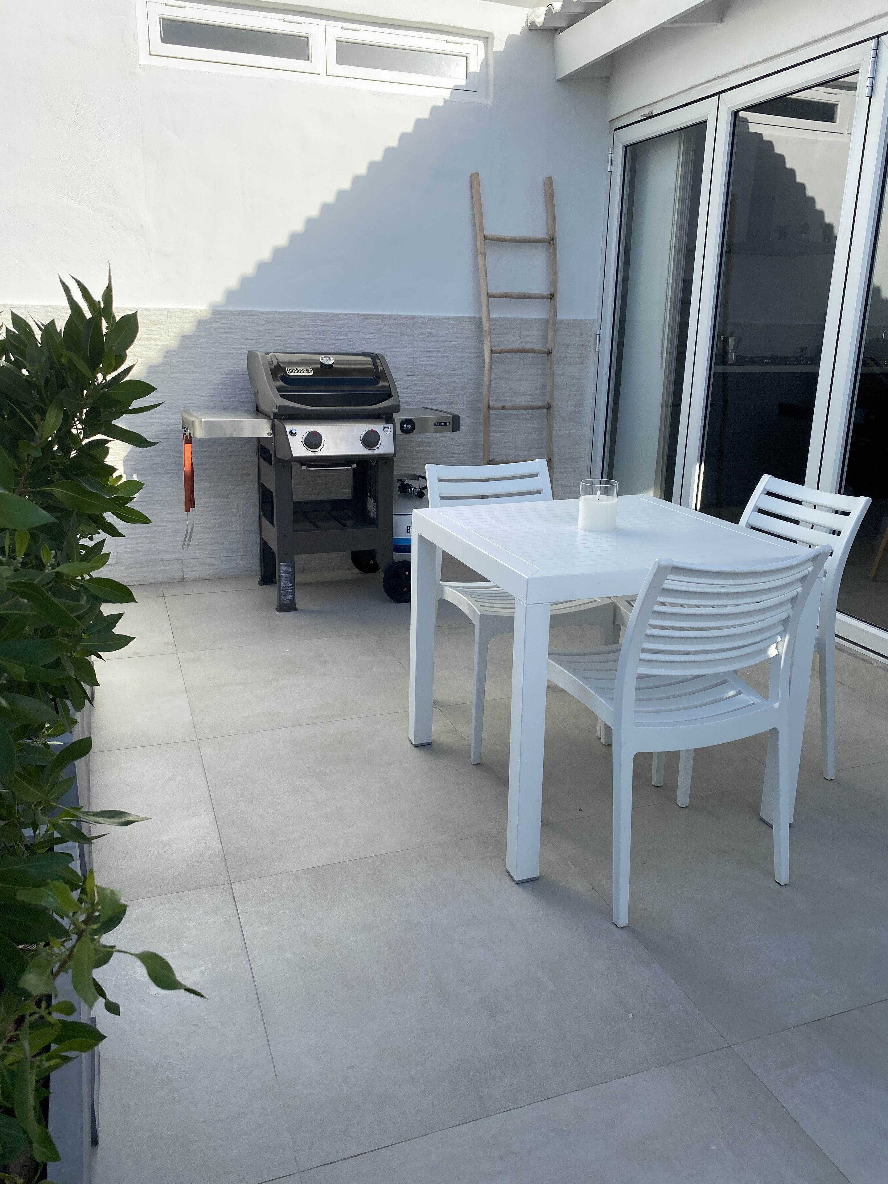 Un patio al aire libre con una mesa y sillas blancas, una parrilla y una puerta corrediza de vidrio.