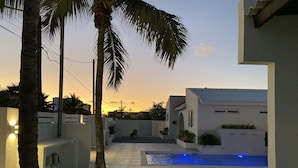 Un área de piscina exterior moderna con cielo despejado, palmeras y un edificio blanco con techo azul.