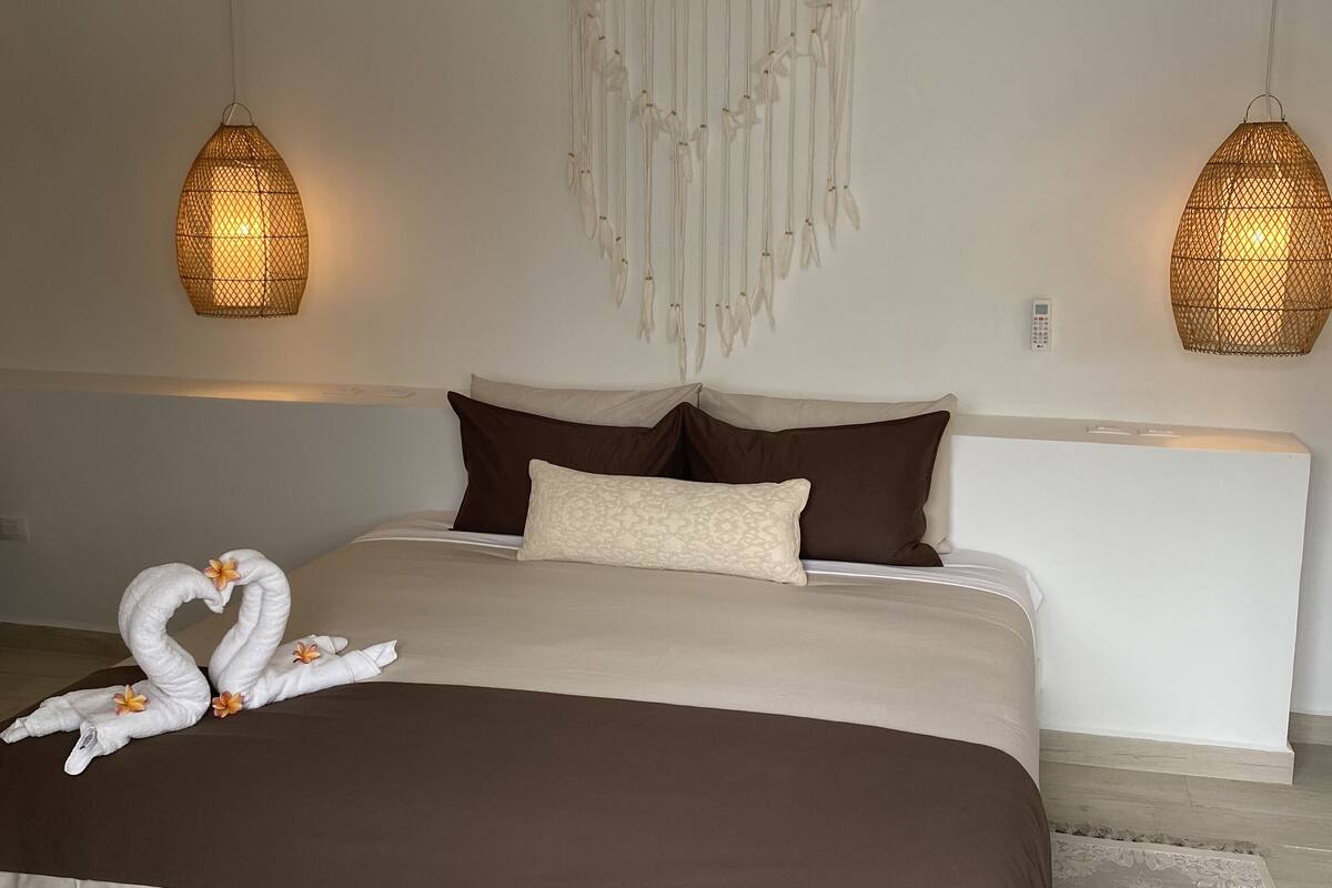 A bedroom with a large bed, two hanging lamps, a dreamcatcher, and swan-shaped pillows.