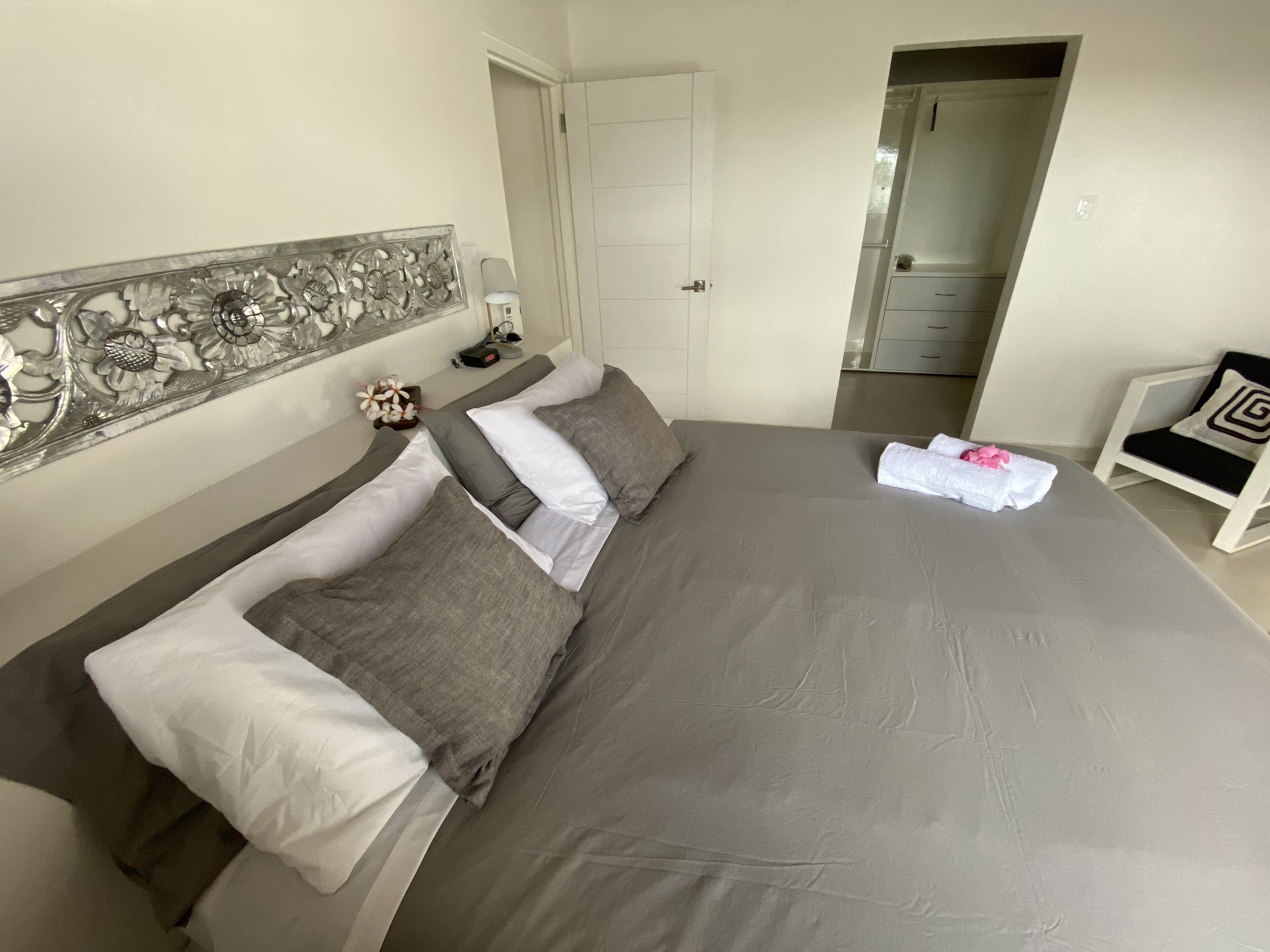 Un lit bien fait, avec une literie grise et des oreillers blancs, un élément décoratif mural, une petite table avec un téléphone et un chargeur, et une chaise avec une serviette.