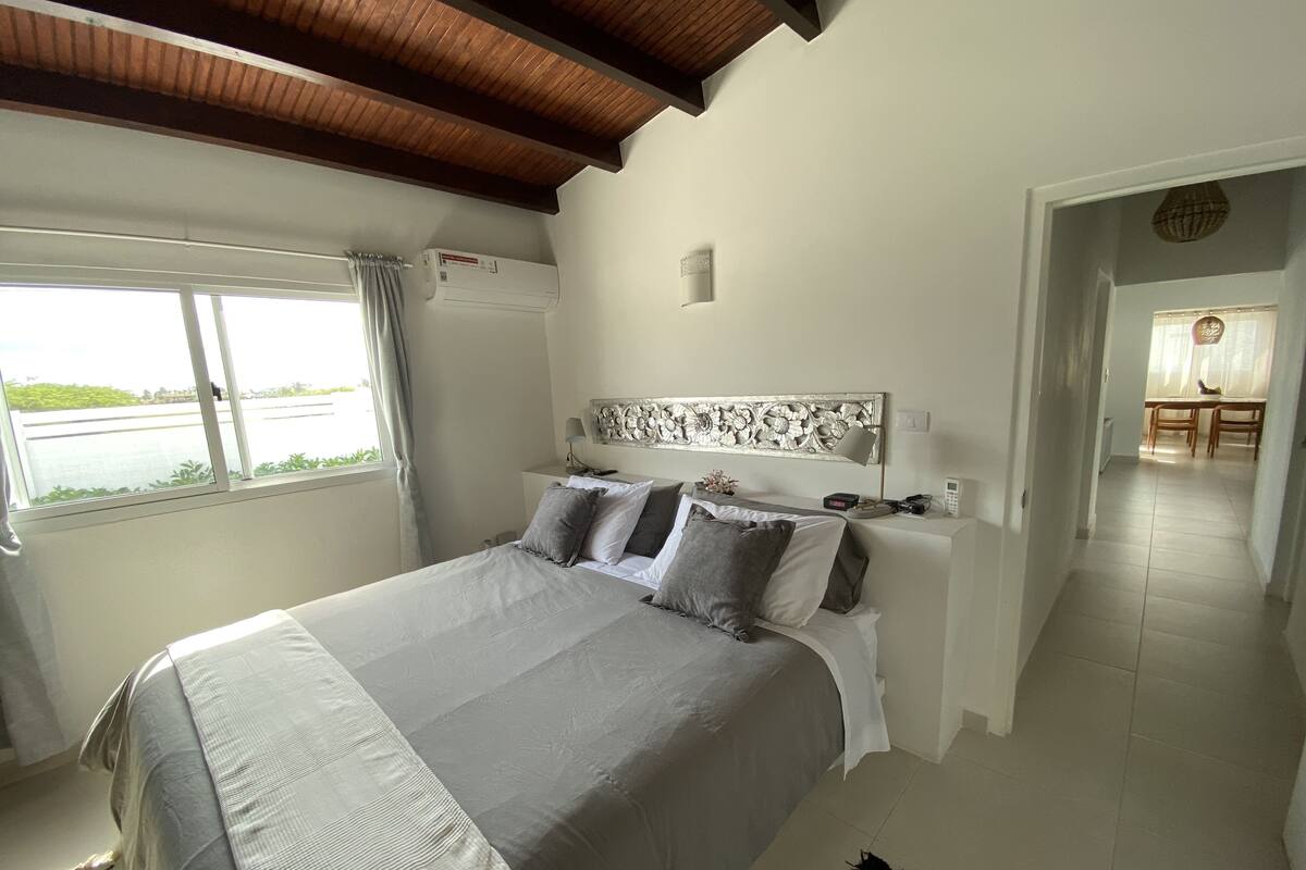 A bedroom with a large bed, a window with curtains, a ceiling fan, and a view of a body of water.