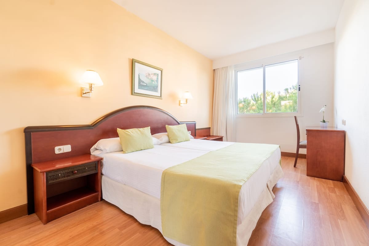 A hotel room with a large bed, two bedside tables, a nightstand, a desk, a chair, and a window with a view of greenery.
