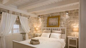 Une chambre avec un mur en pierre, un lit avec une literie blanche, deux tables de chevet avec des lampes et une fenêtre avec des rideaux blancs.